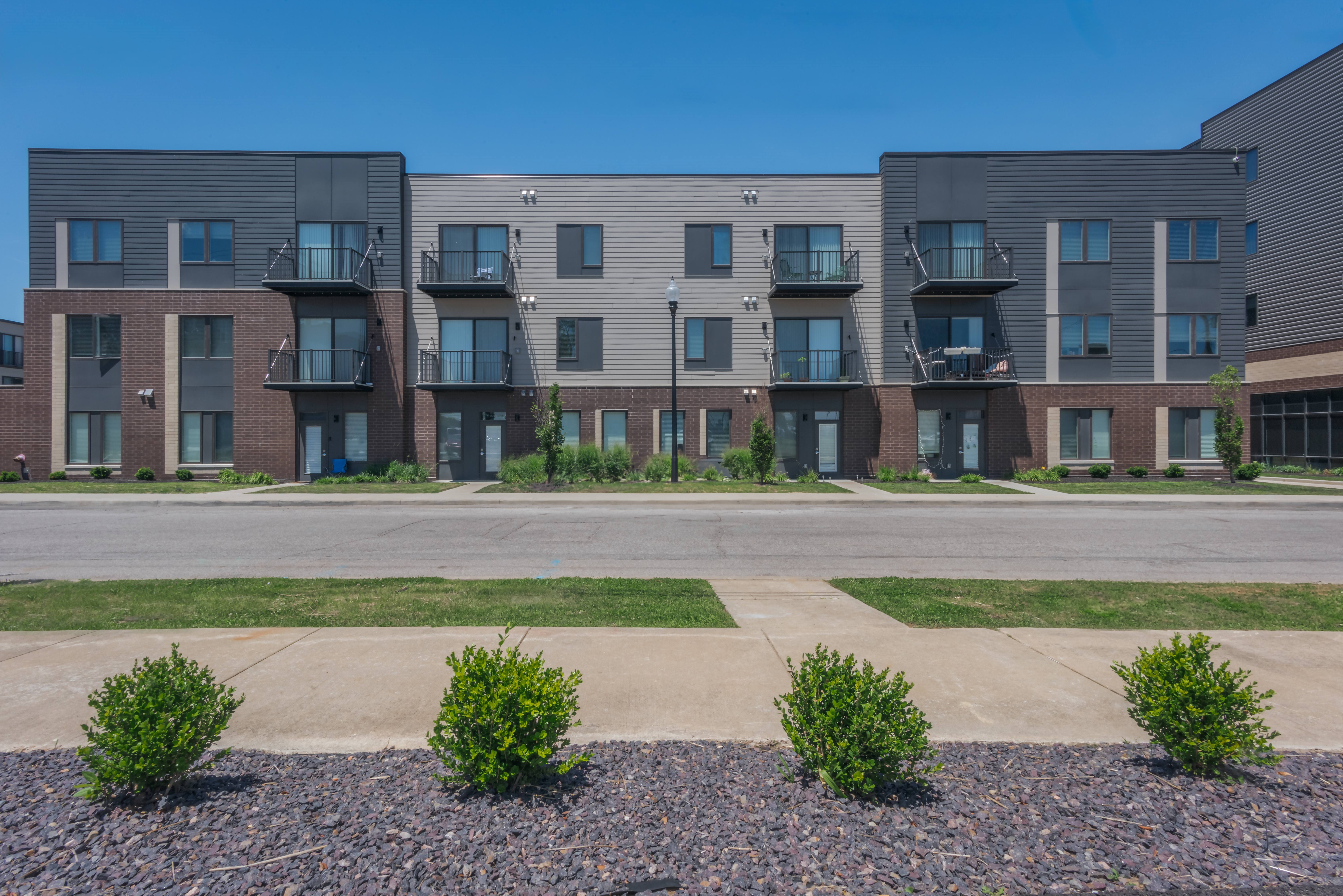 Apartment complex with grey and brown buildings and green bushes in front.